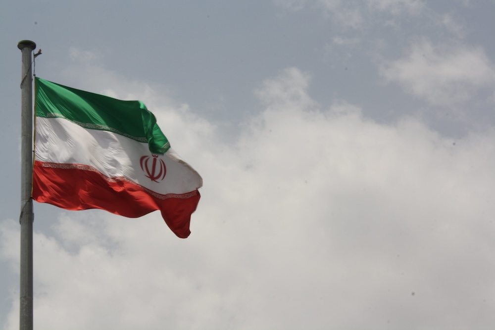 Photograph of the flag of Iran unfurled against a sky with clouds.